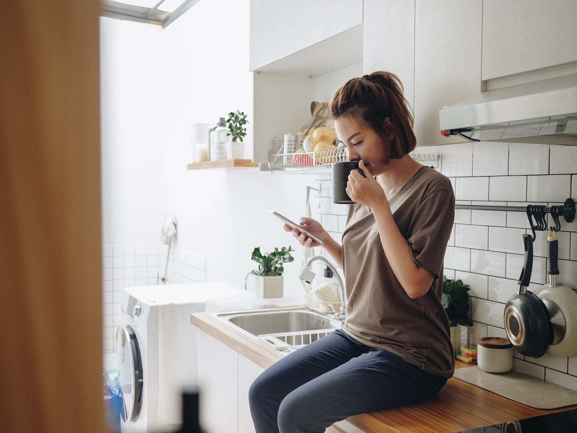 Teenage asian woman drinking coffee while sitting on kitchen counter and working on smart phone in morning at home.